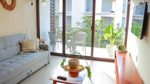 A living room with a sofa, a coffee table with a plant, a TV on the wall, and a balcony with chairs and a potted plant. - Gama 09 Apartments - Apartment with balcony (Tulum)