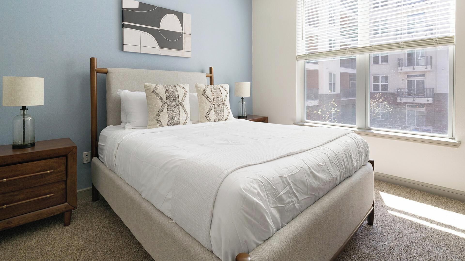 A bedroom with a bed, nightstands, a lamp, a window with blinds, and a city view.