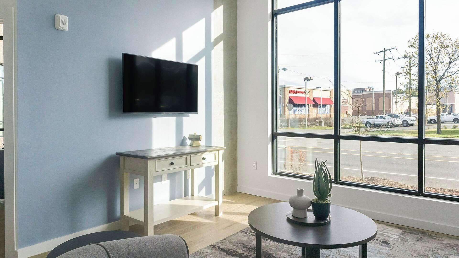 Una habitación con una televisión de pantalla plana montada en la pared, un escritorio pequeño blanco con cajones, una mesa redonda con una planta en maceta y un amplio ventanal con vistas a la calle y edificios.