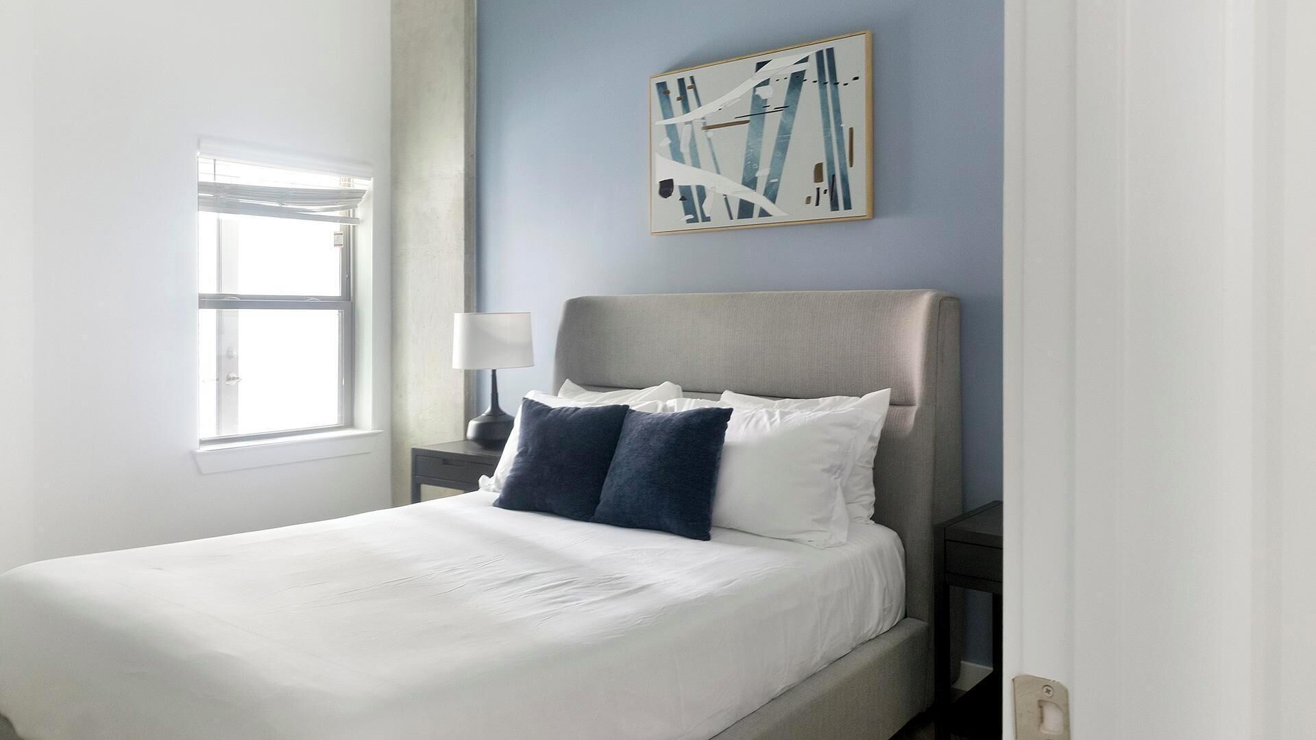 A neatly made bed with white linens and two dark pillows, a bedside table with a lamp, and a wall-mounted abstract art piece.
