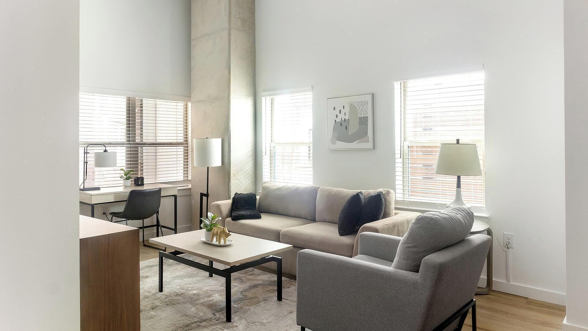 Ein modernes Wohnzimmer mit Sofa, Ohrensessel, Couchtisch und Schreibtisch mit Stuhl. Es gibt Fenster mit Jalousien, eine Lampe und ein gerahmtes Bild an der Wand.