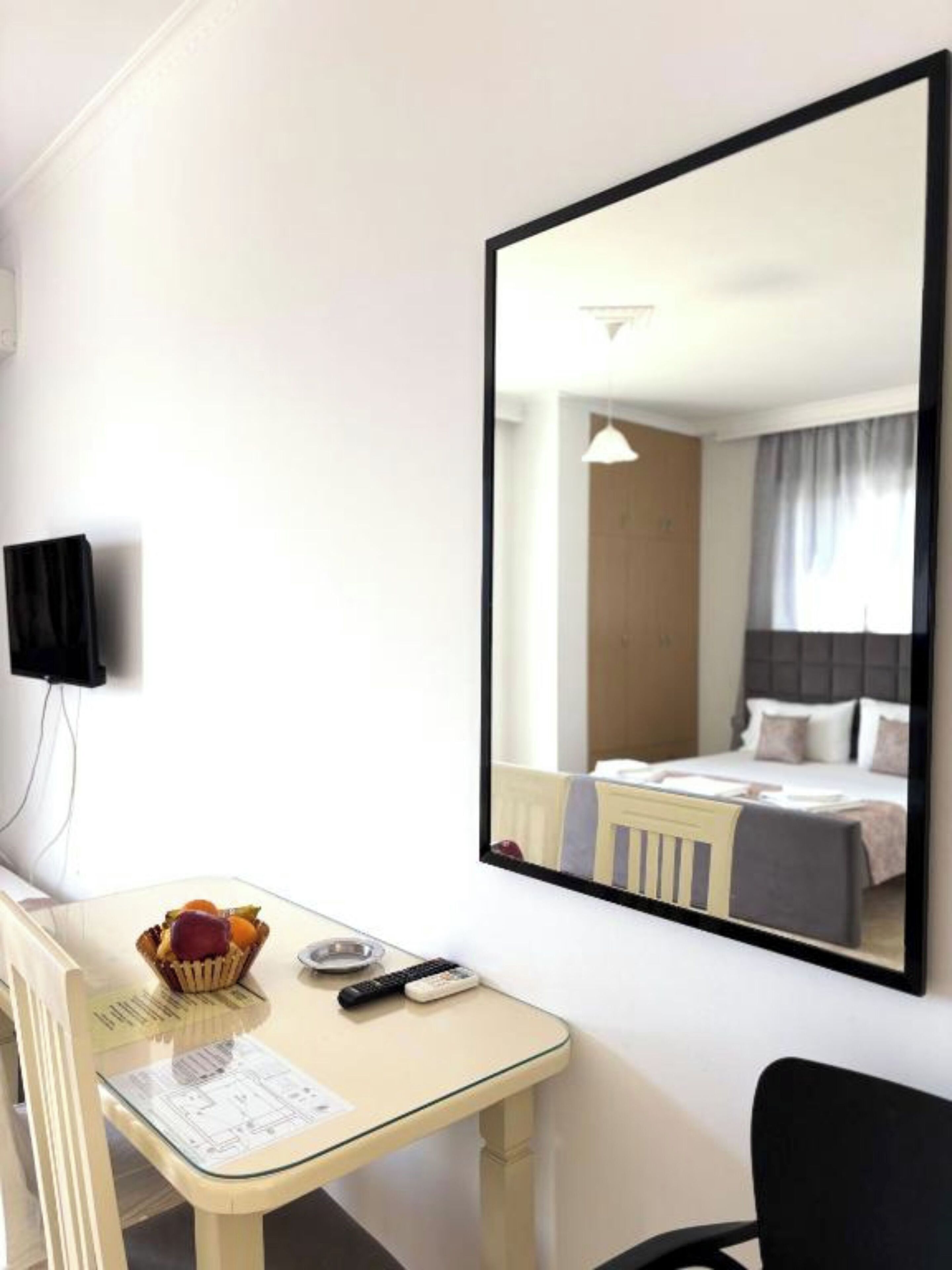 A hotel room with a bed, a desk with a basket of fruit, a remote control, and a mirror reflecting the room.