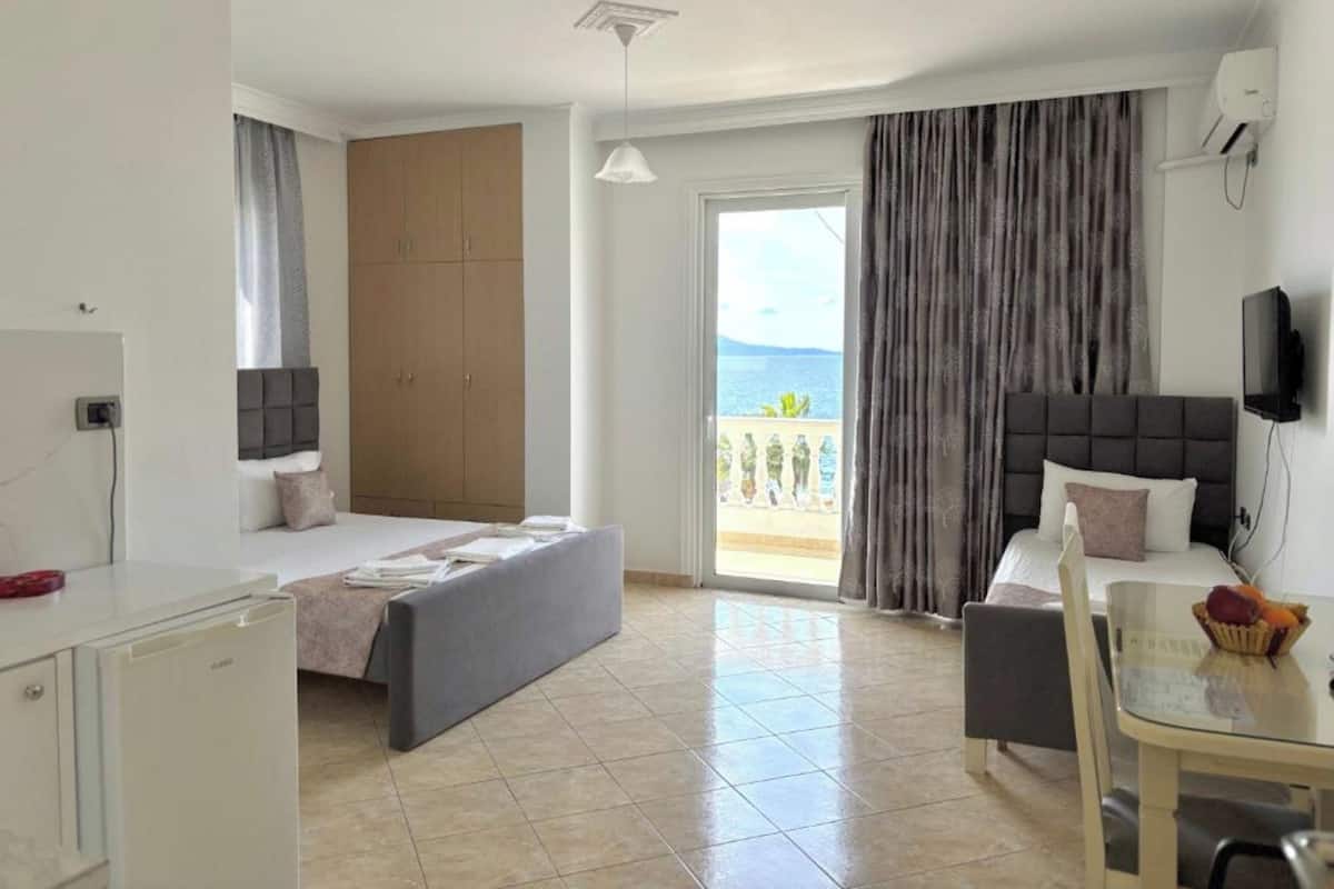 A hotel room with a bed, a desk, a chair, a TV, and a balcony with a view of the sea.