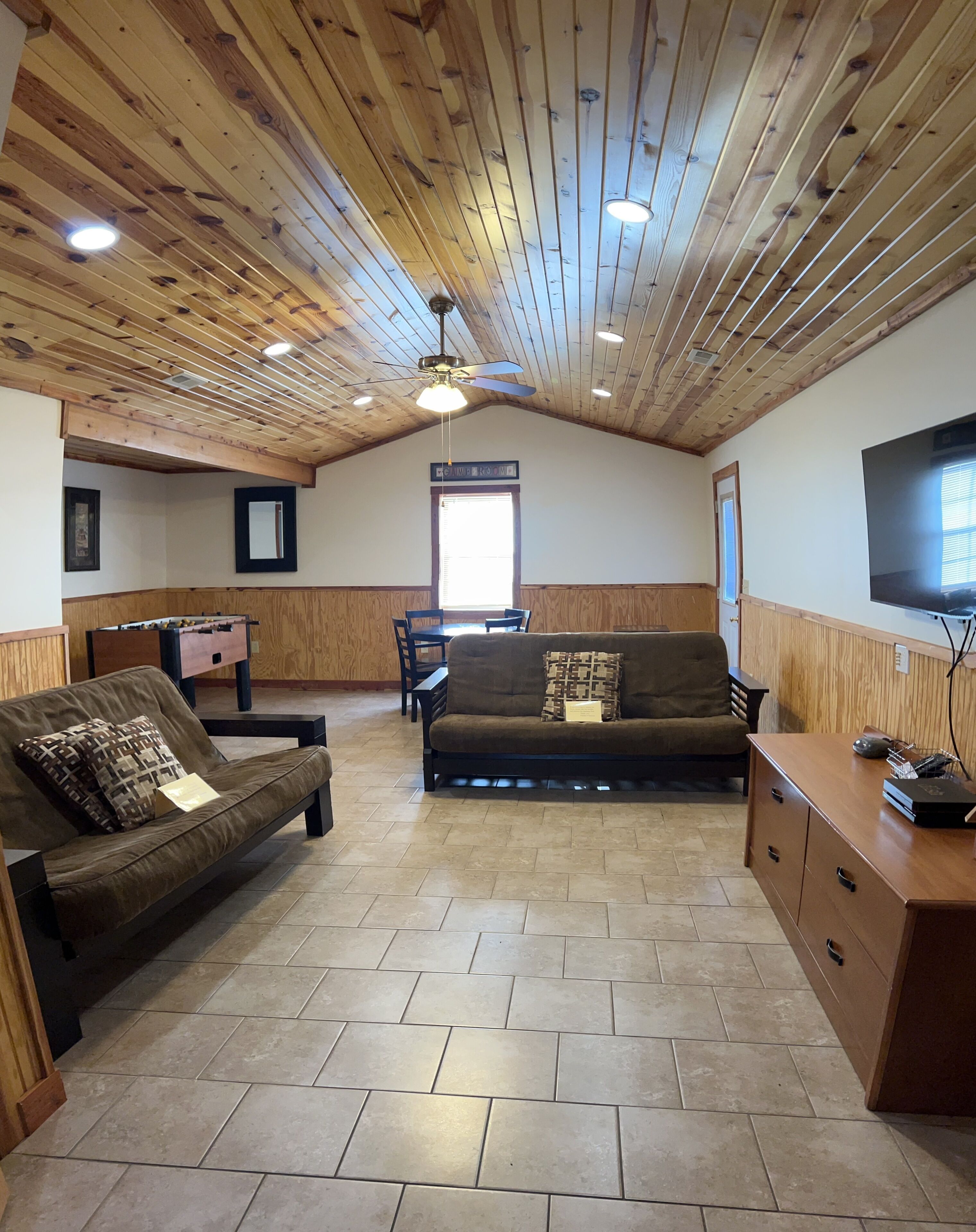 A cozy living room with a wooden ceiling, a sofa, a coffee table, a TV, and a foosball table.