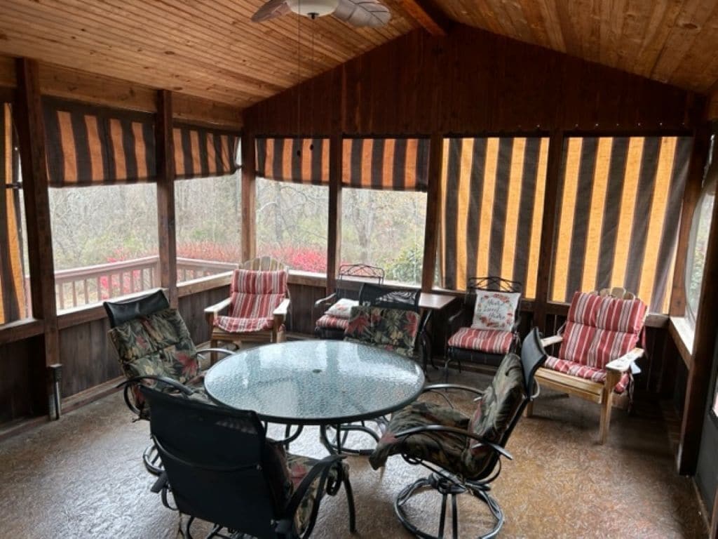 A covered patio with a round glass table surrounded by chairs, large windows with striped curtains, and a wooden ceiling.