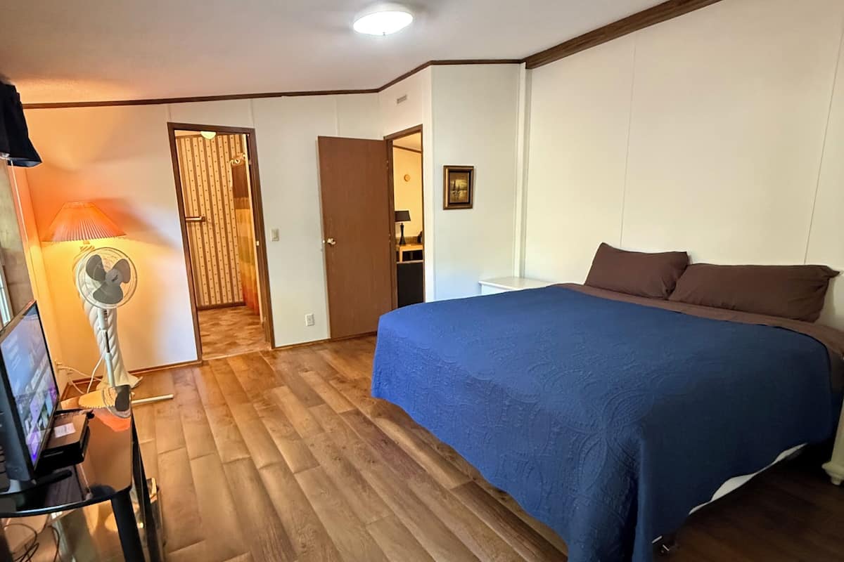 A bedroom with a bed, a desk with a computer, a standing fan, and a door leading to another room.