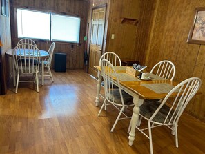 Un coin repas avec une table en bois et des chaises blanches, un petit haut-parleur noir et une fenêtre avec des stores.