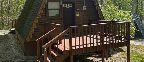 A wooden cabin with a steeply pitched roof, a front door, and a wooden staircase leading to a deck.