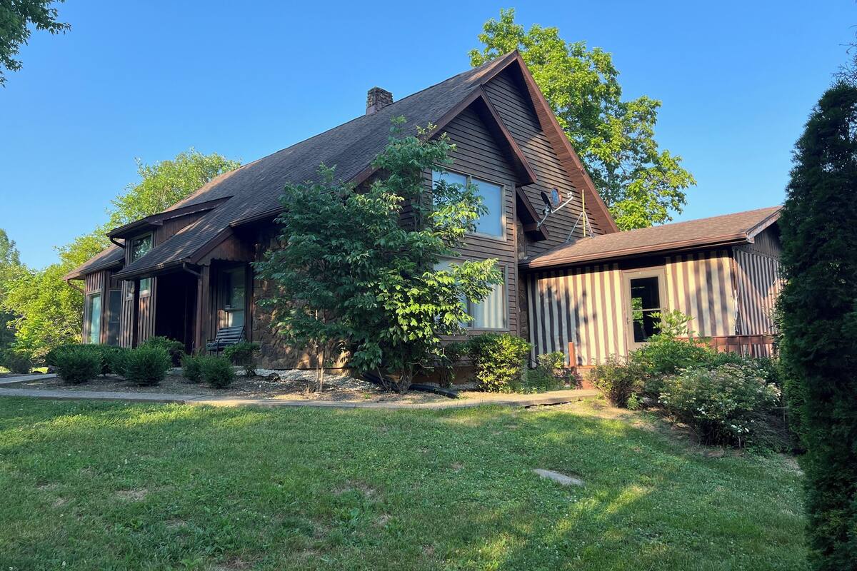 A two-story house with a stone foundation, a sloped roof, and a large front yard with mature trees and well-maintained grass.