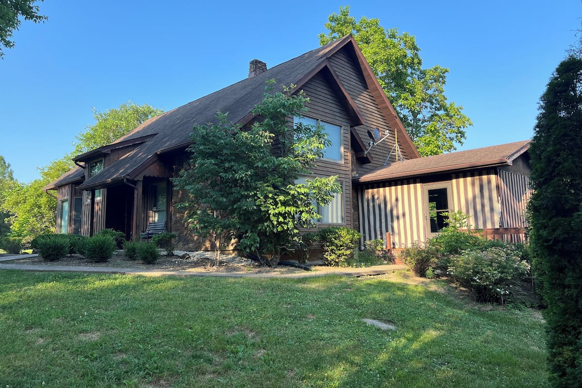 A two-story house with a stone foundation, a sloped roof, and a large front yard with mature trees and well-maintained grass.