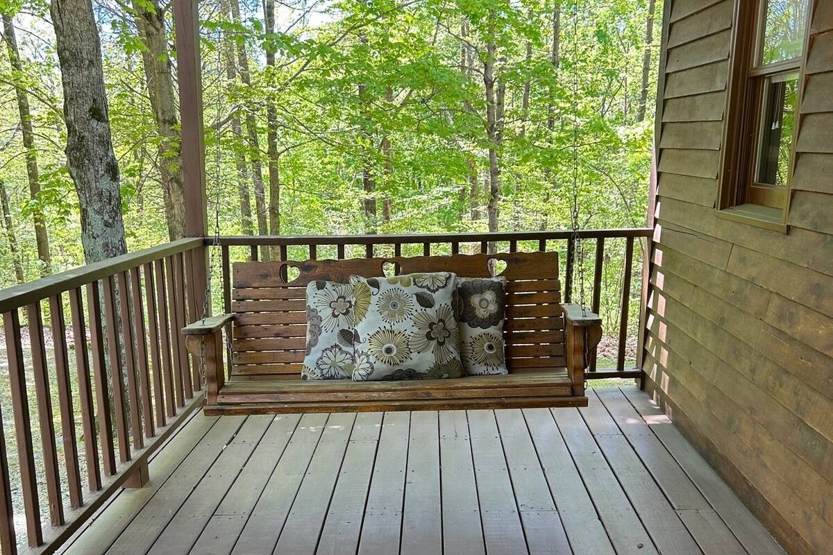 A wooden porch with a swing seat and cushions, surrounded by trees.