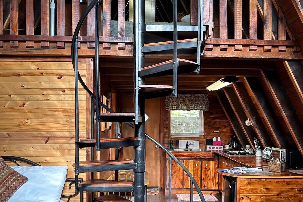 A rustic cabin interior with a spiral staircase leading to a loft bedroom, a kitchen area, and a dining space.