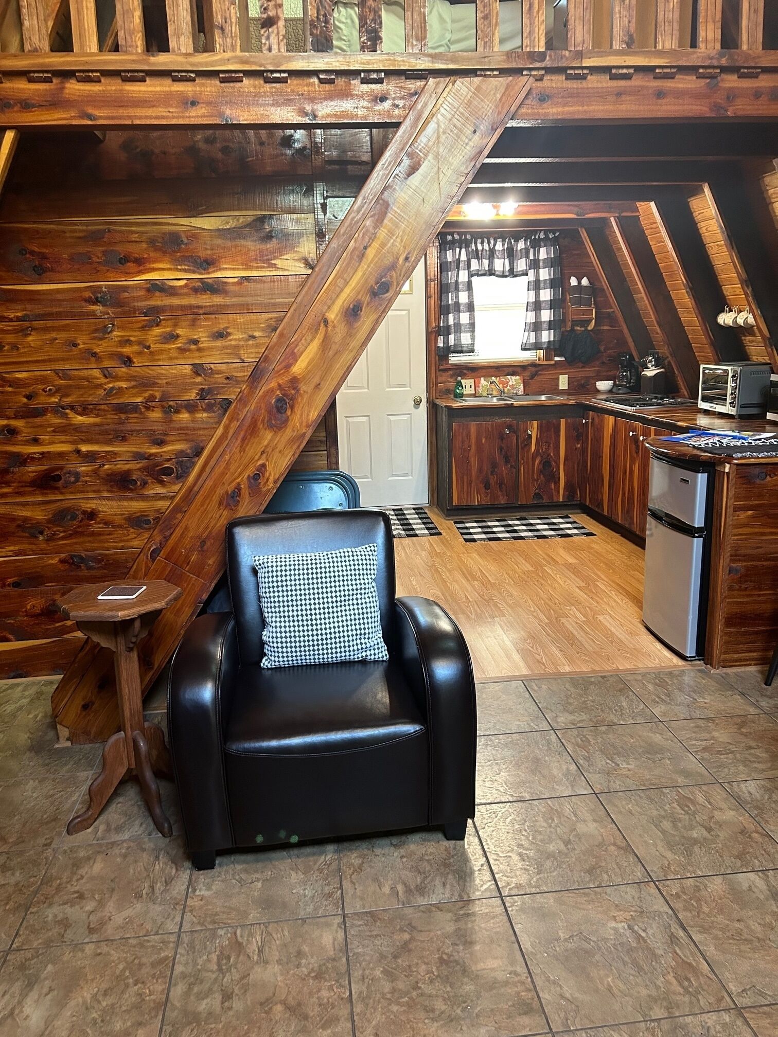 A cozy room with a black leather chair, a wooden staircase, a kitchen area, and a tiled floor.
