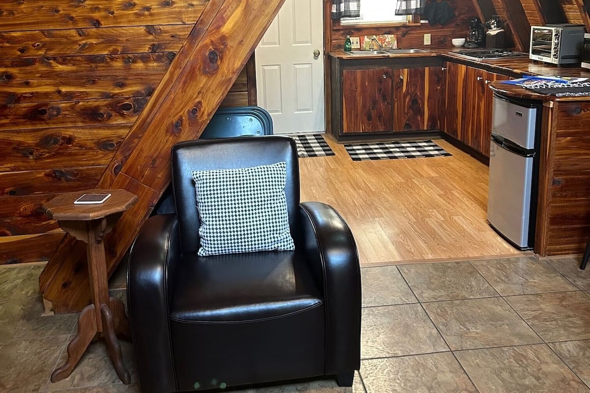 A cozy room with a black leather chair, a wooden staircase, a kitchen area, and a tiled floor.