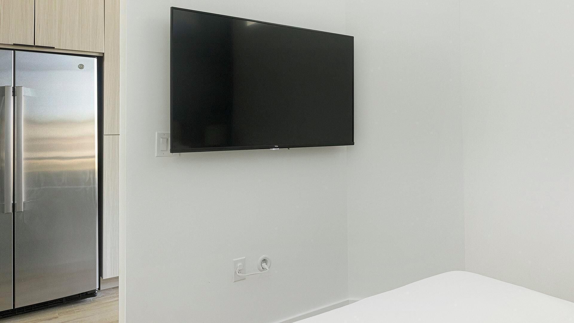 A modern kitchen with a stainless steel refrigerator, a flat-screen TV mounted on the wall, and a white countertop.