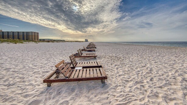 En strand med træliggestole stillet op i rækker, sand og en bygning i det fjerne.