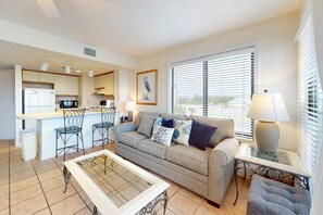 A compact living space with a kitchenette, a sofa, a coffee table, and a window with blinds. - Waterfront condo with pool & hot tub (Fort Morgan)