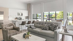 Una sala moderna con un sofá gris, dos sillones, una mesa de centro y una cocina con armarios blancos y un microondas.