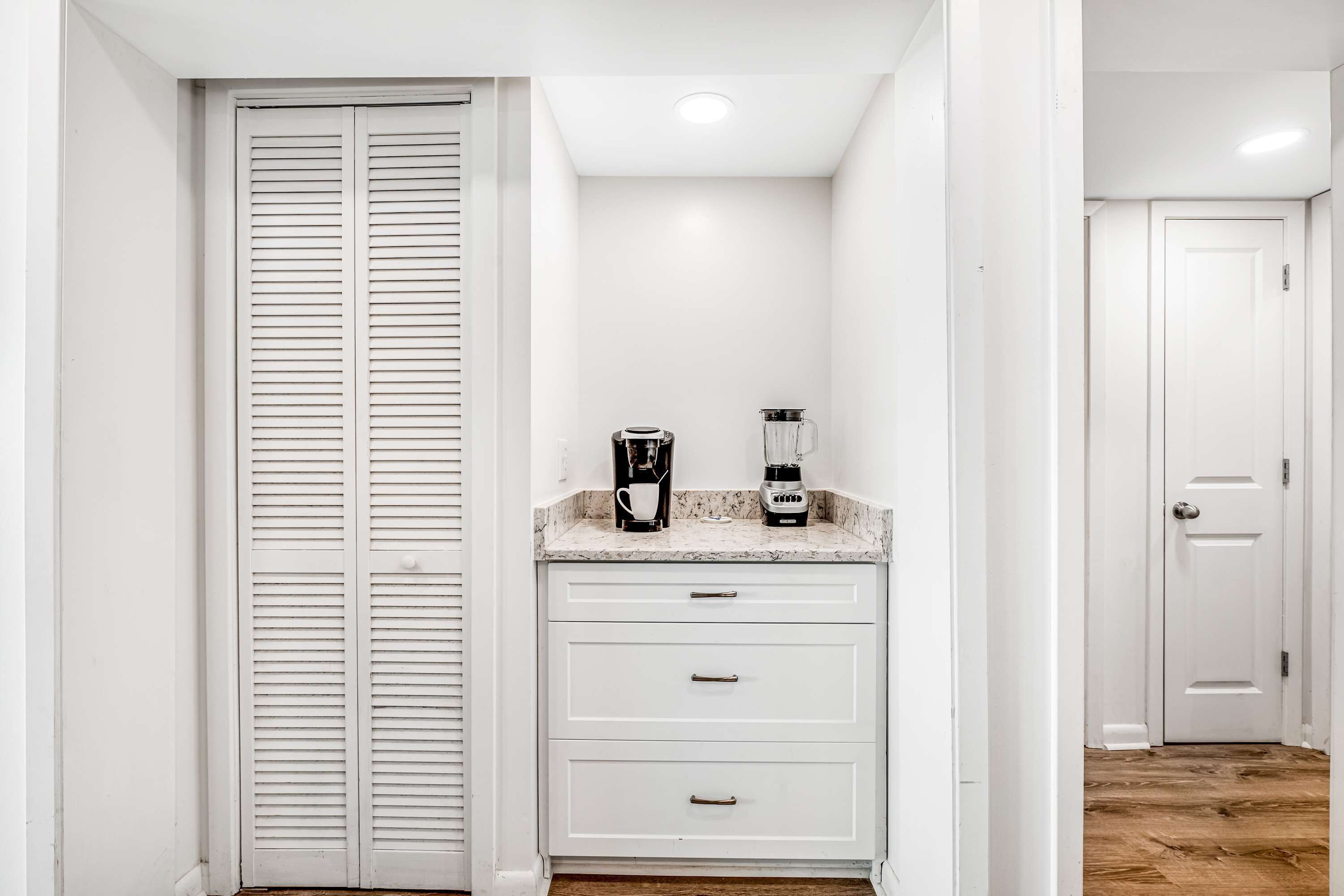A white closet with sliding doors, a countertop with a coffee maker and blender, and a wooden floor.