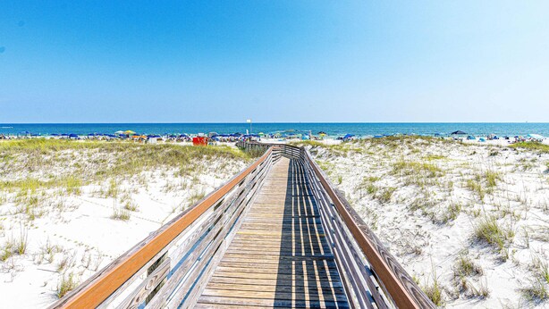 A wooden boardwalk leading to a beach with sand dunes and beach umbrellas. - Waterfront condo with shared pool & hot tub - near dining & the beach (Fort Morgan)
