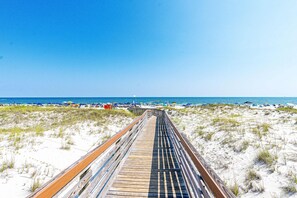 A wooden boardwalk leading to a beach with sand dunes and beach umbrellas. - Waterfront condo with shared pool & hot tub - near dining & the beach (Fort Morgan)