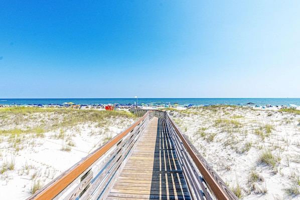 Ein hölzerner Steg führt zu einem Strand mit Dünen und Strandsonnenschirmen.
