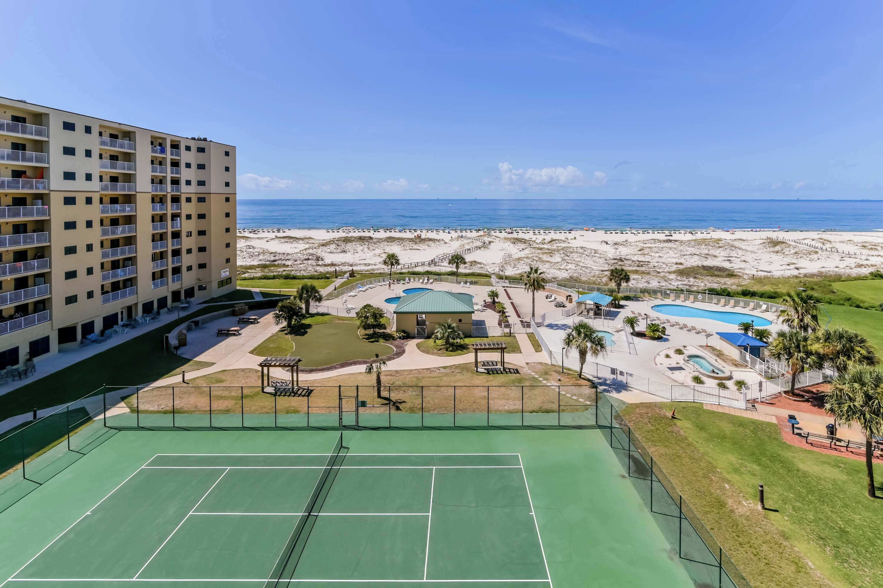 Un hotel de varios pisos con cancha de tenis, zona de piscina y vista al mar.
