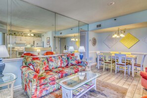 Ein Wohnzimmer mit einer Couch, einem Glastisch, einem Essbereich mit Stühlen und einer Küche im Hintergrund.