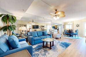 A spacious living room with a blue sofa, a glass coffee table, and a ceiling fan. - Plantation Dunes condo with Gulf views, pool, hot tubs & gym - walk to beach (Fort Morgan)