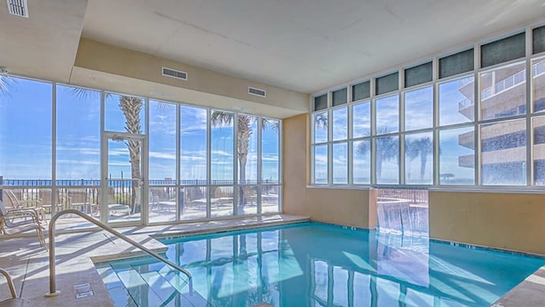An indoor swimming pool with clear windows, a poolside area, and a view of palm trees. - Waterfront home with a private deck, gas grill, WiFi, & central AC (Gulf Shores)