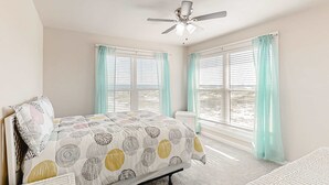 Una camera da letto con un letto, una ventola a soffitto, due finestre con tende e vista sulla spiaggia.