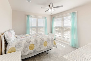 Ein Schlafzimmer mit einem Bett, einem Deckenventilator, zwei Fenstern mit Vorhängen und Blick auf den Strand.