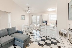 Ein modernes Wohnzimmer mit grauem Sofa, einem Essbereich mit weißen Stühlen, einer Küche mit weißen Schränken und einem Schachbrettfußboden.