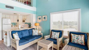 A living area with wicker furniture, a kitchen in the background, and a window with blinds. - Third-floor condo with balcony, close beach access, pools, hot tubs & tennis (Fort Morgan)