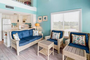 A living area with wicker furniture, a kitchen in the background, and a window with blinds.