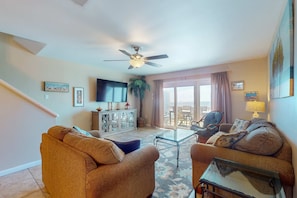 A living room with a ceiling fan, a television, a sofa, a coffee table, and a view through a glass door. - Gulf-Front: Dolphins, Starlight & Steps to Beach (Fort Morgan)