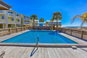 A wooden deck with a swimming pool, surrounded by multi-story buildings with balconies and palm trees. - Gulf-Front: Dolphins, Starlight & Steps to Beach (Fort Morgan)