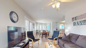 Ein Wohnzimmer mit einer Couch, Sesseln, einem Fernseher, einem Holztisch und einem Essbereich mit Tisch und Stühlen.