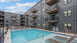 Un moderno edificio de apartamentos con piscina en la azotea, varios balcones y un cielo azul despejado.