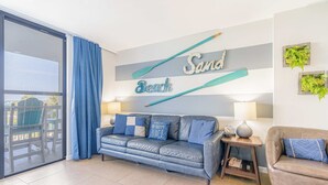 Uma sala de estar com um sofá azul, uma poltrona marrom, uma porta de vidro que dá para uma varanda e uma parede com a inscrição 'Sand Beach' e um morcego.