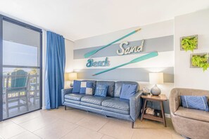 A living room with a blue sofa, a brown armchair, a glass door leading to a balcony, and a wall with the words 'Sand Beach' and a bat. - Spacious condo with pool & hot tub - tennis available on-site (Gulf Shores)
