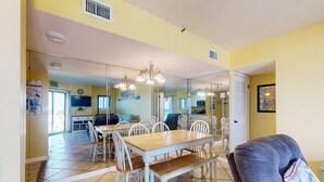 A dining area with a wooden table and white chairs, a mirror reflecting the room, and a gray sofa. - Snowbird-friendly condo with WiFi, on-site pools, hot tub, golf, & tennis (Gulf Shores)