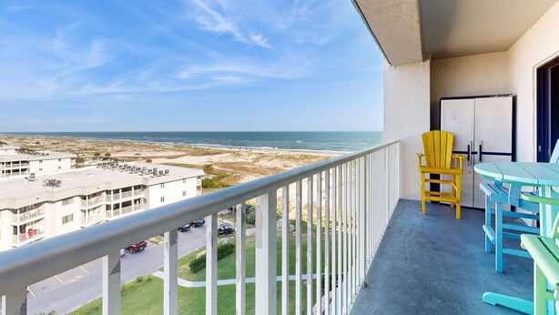 A balcony with a view of the ocean, a table, chairs, and a refrigerator. - Snowbird-friendly condo with WiFi, on-site pools, hot tub, golf, & tennis (Gulf Shores)