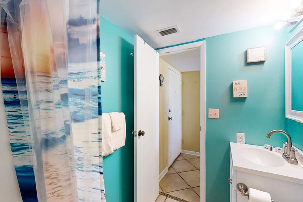 A bathroom with a white sink, a shower curtain with a beach scene, and a door leading to a hallway. - Snowbird-friendly condo with WiFi, on-site pools, hot tub, golf, & tennis (Gulf Shores)