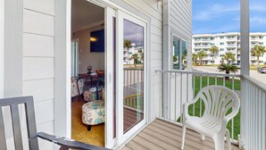 A balcony with a white plastic chair, a wooden bench, and a sliding glass door leading to an interior room with a patterned ottoman and a table. - Spacious condo with balcony and shared pools and tennis, on-site golfing (Gulf Shores)