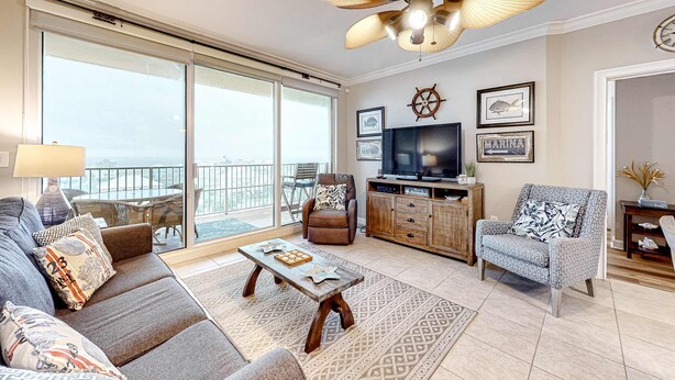 A living room with a sofa, armchair, coffee table, TV, and a balcony with a view. - Updated 5th-floor condo with Gulf view, balcony, pool, & hot tub - near beach (Fort Morgan)