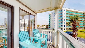 A balcony with two chairs, a palm tree, and a view of a multi-story building with balconies.