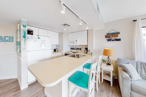 A modern kitchen with a central island, white cabinetry, and a dining area with a striped sofa and teal chairs. - Second-floor condo with pool, hot tub, ocean view, fast WiFi & central AC (Fort Morgan)
