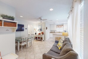 A modern living room with a grey sofa, dining table, and chairs, a kitchen area with white cabinets, and a ceiling fan. - Fun Seahorse condo with gas grill, W&D, & pool access - walk to West Beach (Gulf Shores)