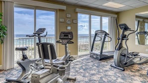 Un gimnasio con bicicletas estáticas y cintas de correr, amplias ventanas con vista al océano y suelo alfombrado.